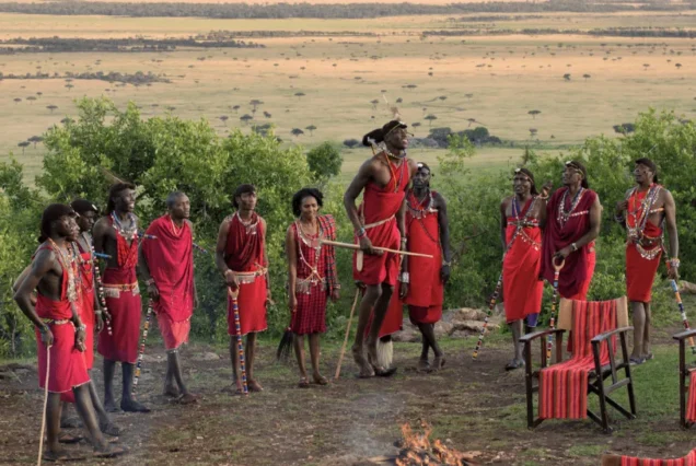 3 Days Tanzania Safari. A group of people in traditional attire gather around a small fire on a grassy area with a scenic landscape in the background. Some are standing and some are seated on chairs adorned with red fabric, enjoying their 2 Nights Tanzania Safari and Adventure Tour. The sky is partially cloudy, giving a peaceful ambiance.